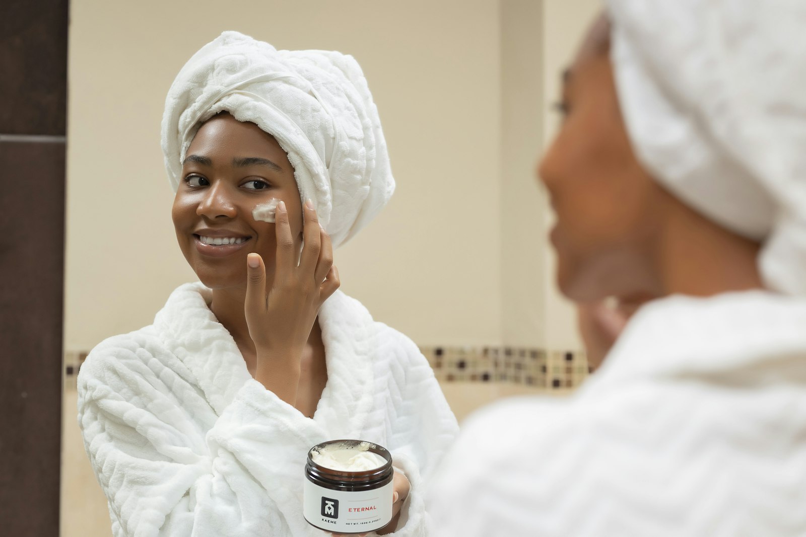 Smiling woman in a white robe and towel applying moisturiser in front of a mirror